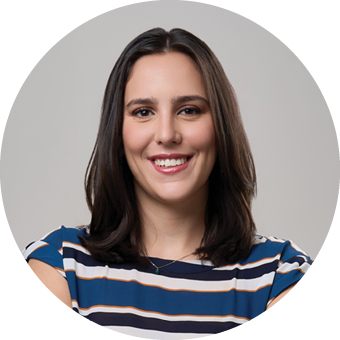 A person with shoulder-length dark hair is smiling and wearing a blue, white, and orange striped shirt against a plain gray background. Description generated by AI