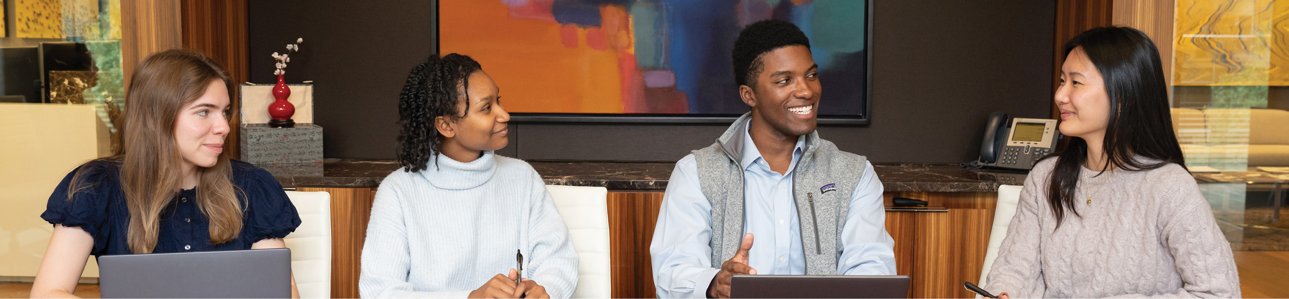 Four adults sit at a table engaged in conversation, with two using laptops and one holding a pen, in a warmly lit room with abstract art on the wall. Description generated by AI